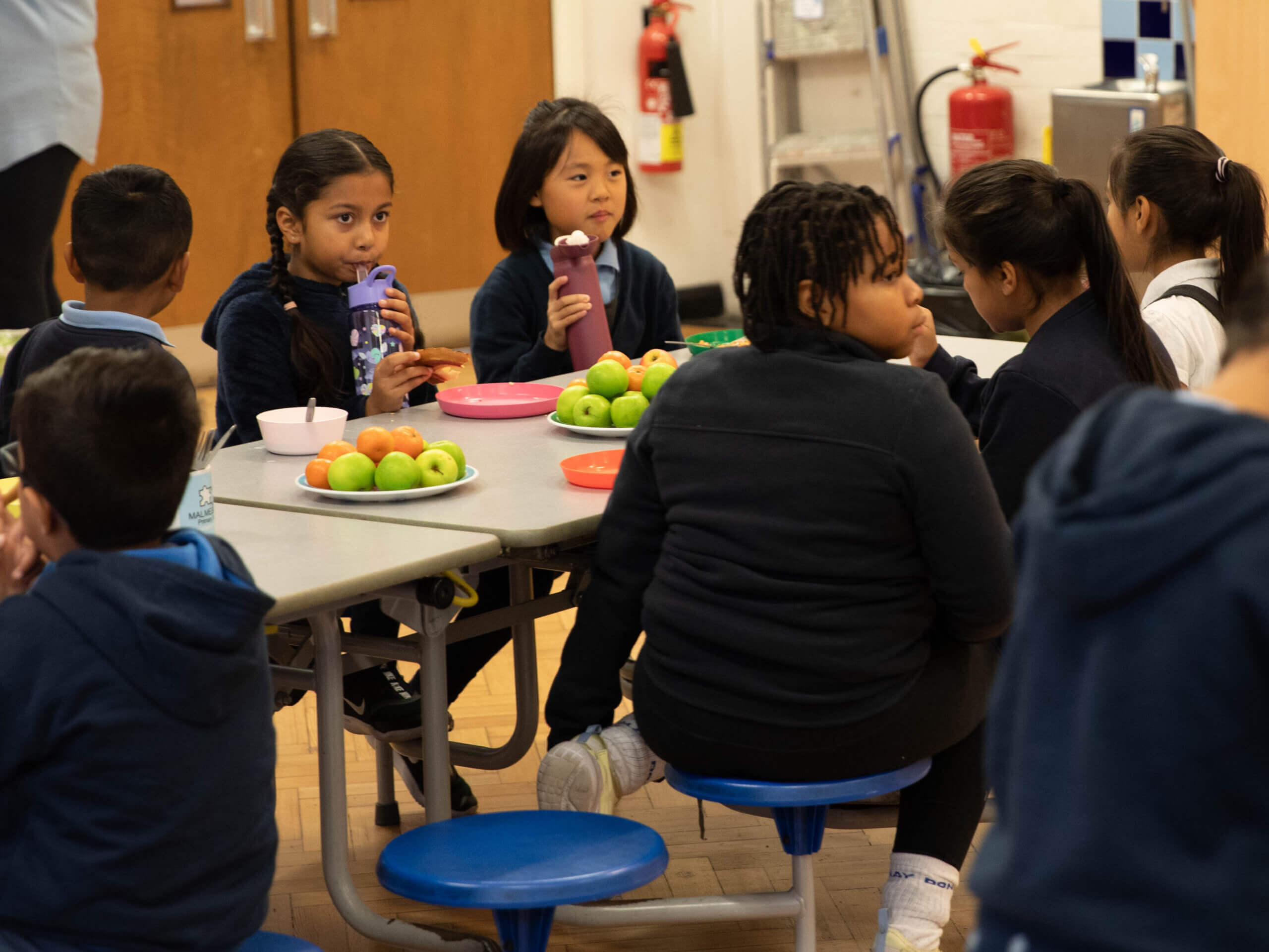 Multiple school children at the breakfast table, drinking water. There is toast and bowls of fruit on the table